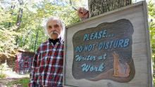 Blei on his own turf in Ellison Bay, Door County, standing in front of a sign welcoming—or, considering the coyote, possibly warning away—visitors to his converted chicken coop writer’s studio. Photo Credit: John Nelson