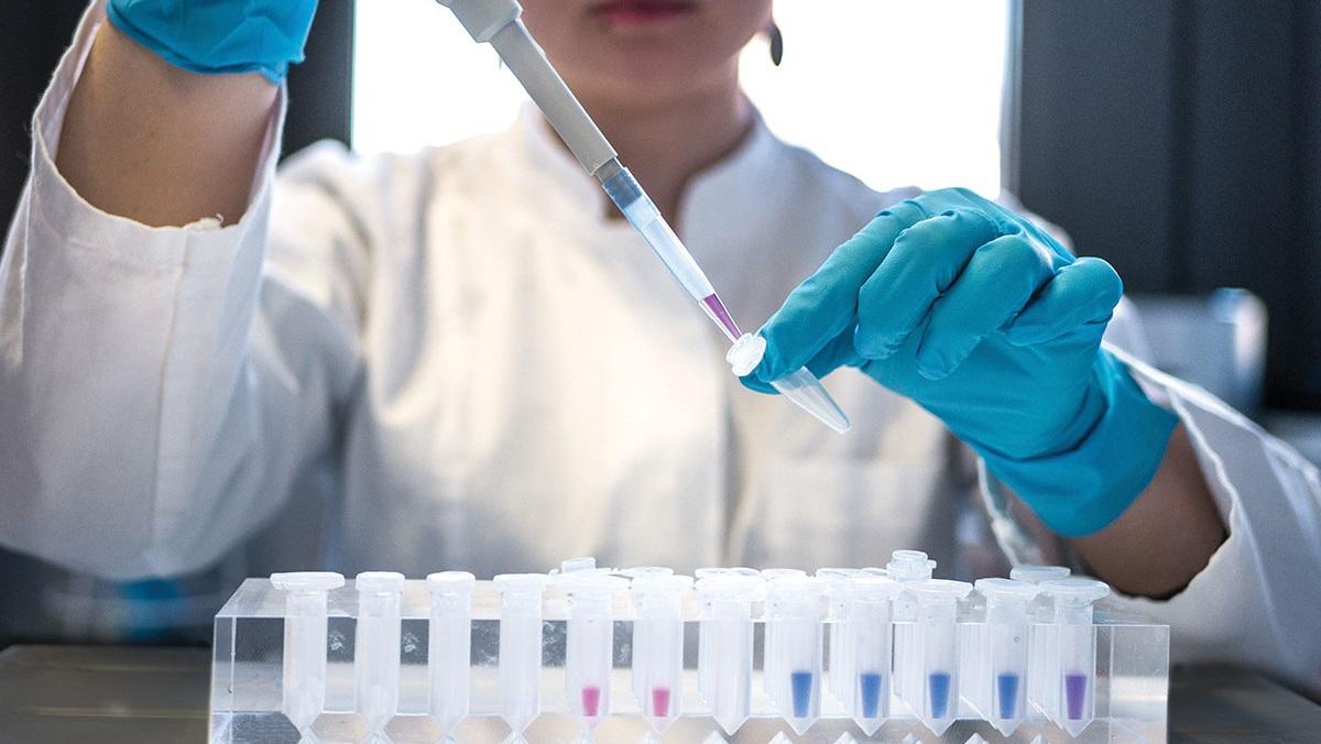 A scientist in a lab coat and gloves uses a pipette