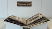 View of open artist book on table with hanging sign behind which reads "Please touch gently" in art gallery setting